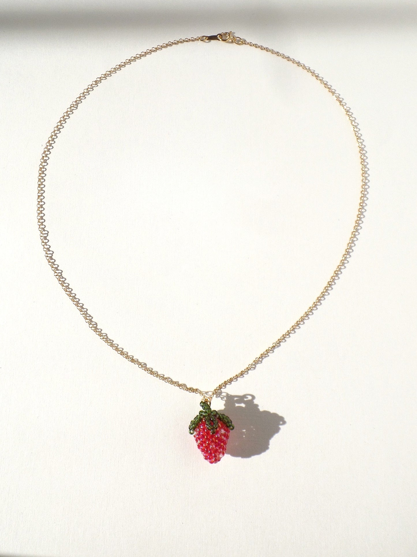 A necklace with a dark red strawberry charm attached to a gold-colored chain, displayed against a white background.