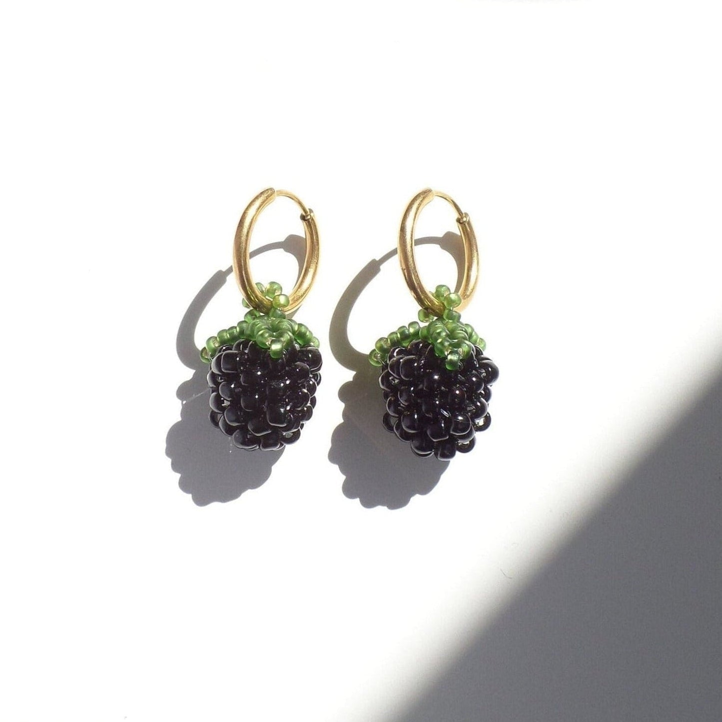 A pair of handwoven blackberry earrings with glass beads, featuring gold-colored hoops, displayed against a shadowy background.